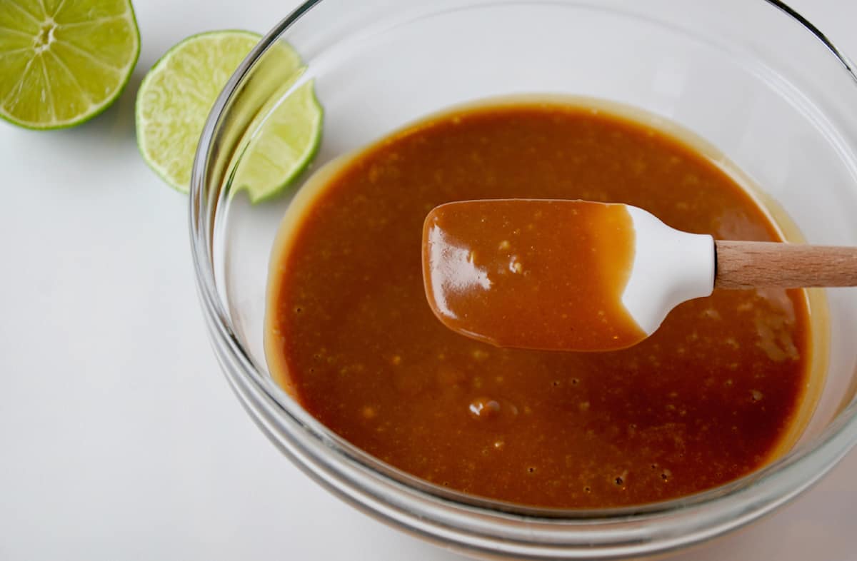 A small silicone spatula in a bowl of creamy peanut sauce. A halved lime sits beside the bowl.