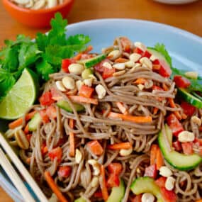 Soba noodle salad topped with peanuts and cilantro in a dinner bowl. Chopsticks sit on the edge of the bowl and small bowls containing more peanuts and quartered limes sit behind it.