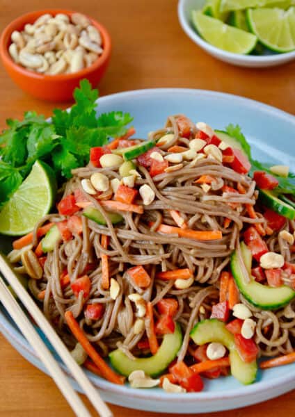 Soba noodle salad topped with peanuts and cilantro in a dinner bowl. Chopsticks sit on the edge of the bowl and small bowls containing more peanuts and quartered limes sit behind it.