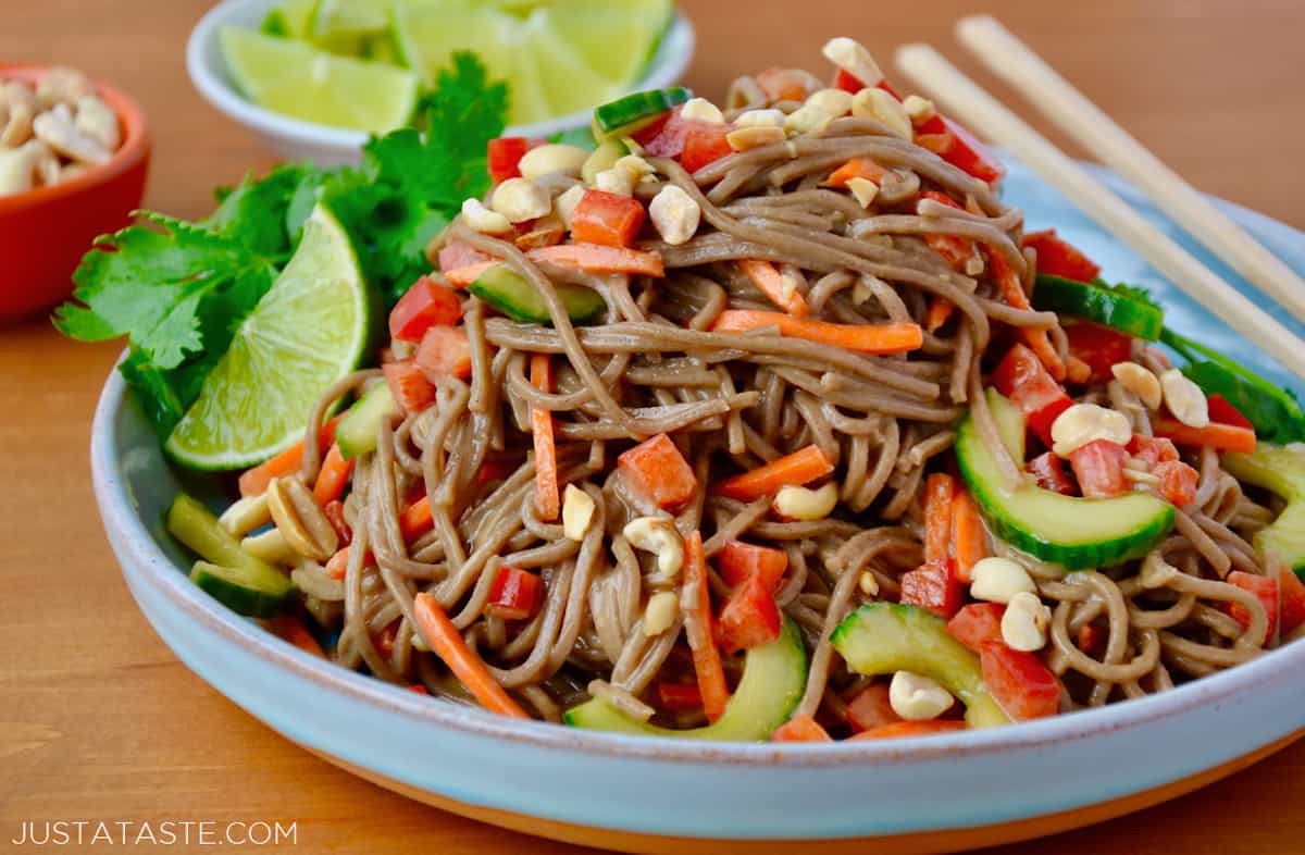 Soba noodle salad topped with peanuts and cilantro in a wide dinner bowl. Chopsticks sit on the edge of the bowl and a quartered lime is on the side.