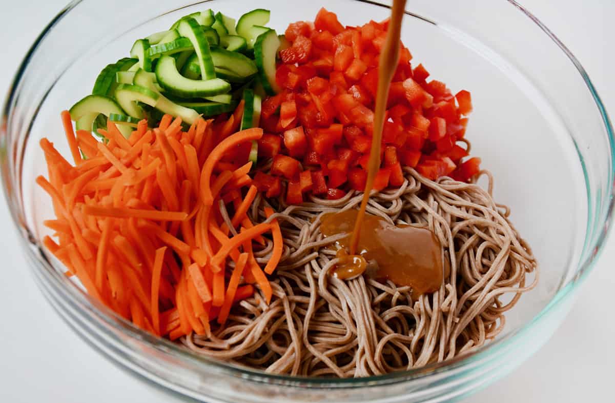Peanut dressing being poured onto cooked soba noodles, diced red bell pepper, sliced cucumber and julienned carrots in a large glass mixing bowl.