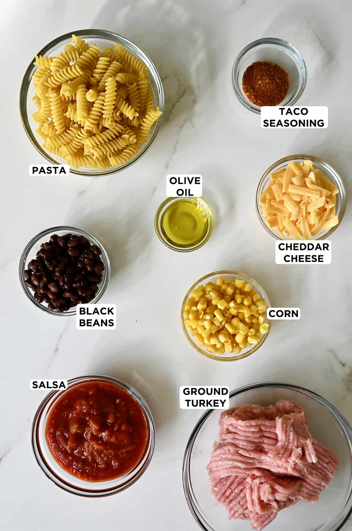 Glass bowls containing the ingredients for turkey taco pasta, including rotini pasta, taco seasoning, shredded cheddar cheese, corn, olive oil, ground turkey, black beans and salsa.