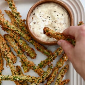 Baked asparagus fries are piled in an oblong white platter, along with a bowl of roasted garlic aioli. A hand is dipping an asparagus fry into the aioli.