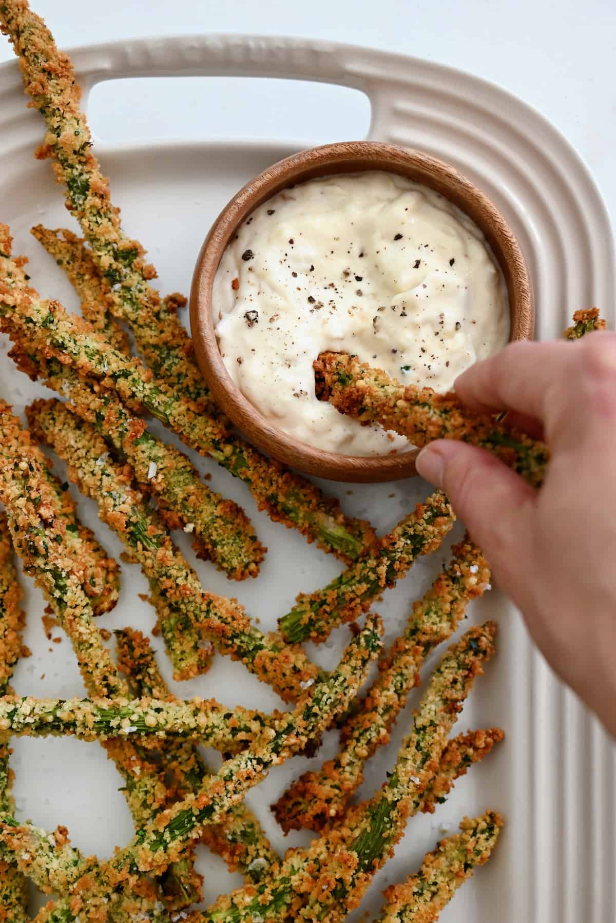 Baked asparagus fries are piled in an oblong white platter, along with a bowl of roasted garlic aioli. A hand is dipping an asparagus fry into the aioli.