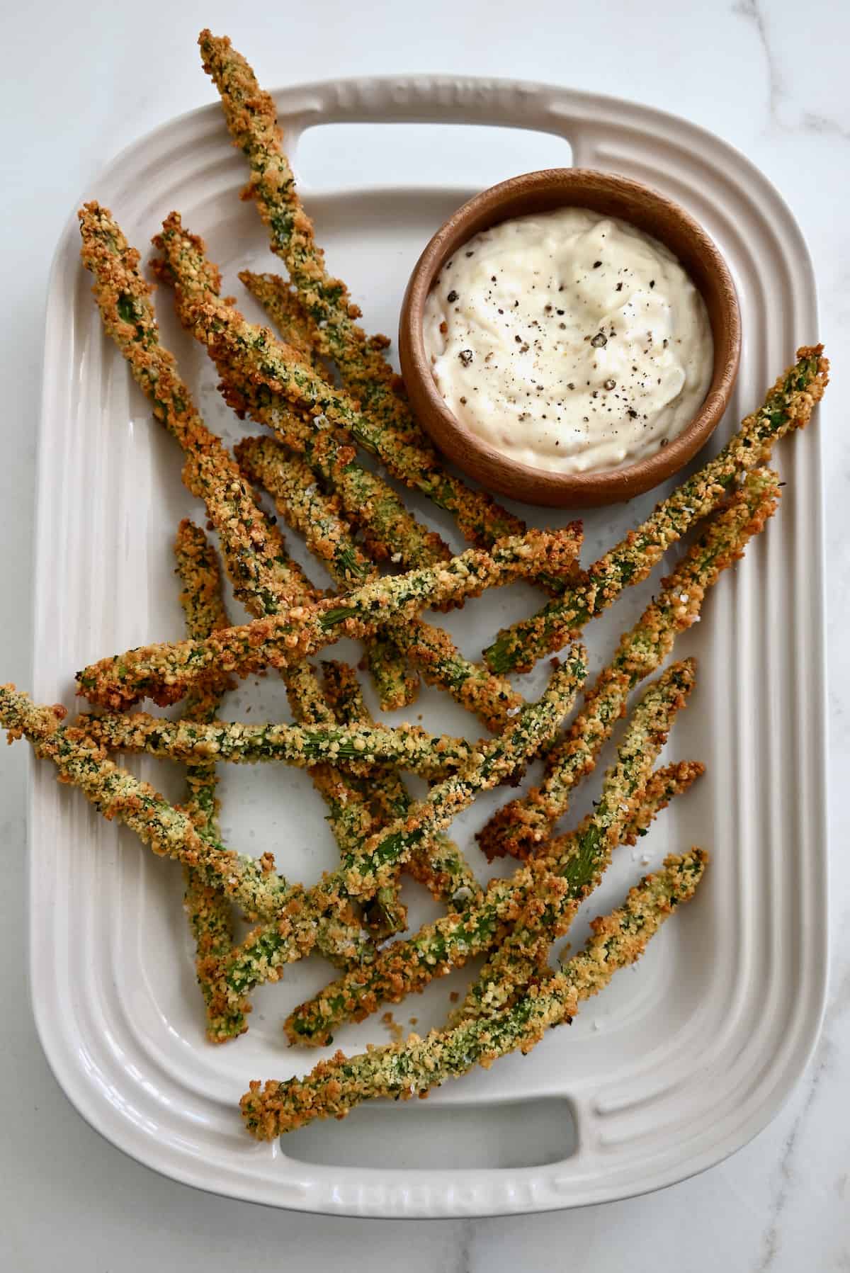 Baked asparagus fries are piled in an oblong white platter, along with a bowl of roasted garlic aioli.
