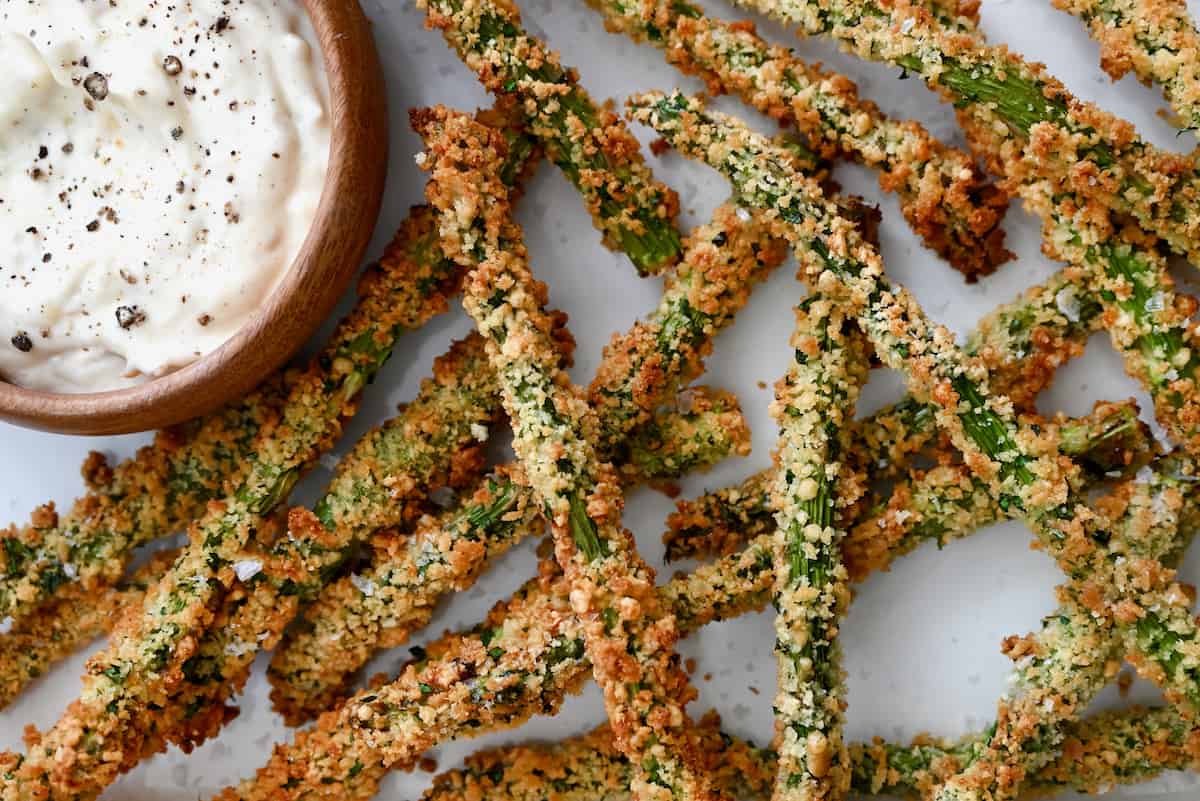 Baked asparagus fries are piled on a white surface, with a bowl of roasted garlic aioli nearby.