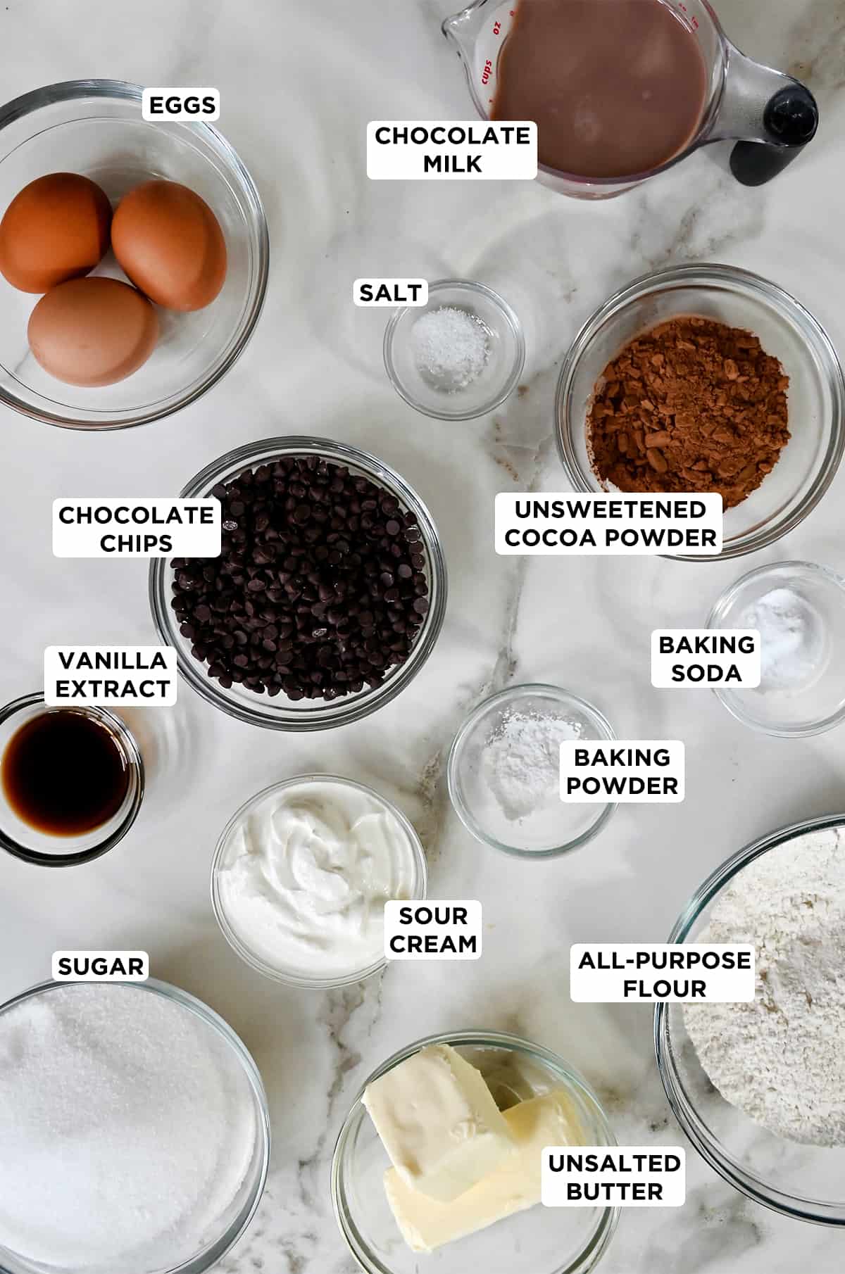 Various sizes of glass bowls containing the ingredients needed to make chocolate cupcakes, including three eggs, chocolate milk, salt, unsweetened cocoa powder, baking soda, baking powder, all-purpose flour, sour cream, chocolate chips, vanilla extract, sugar and unsalted butter.