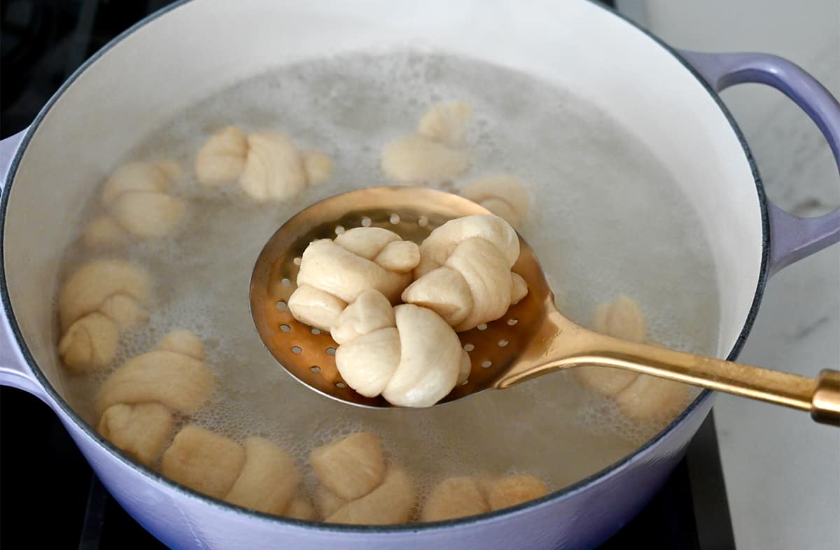 Boiled pretzel knots on a slotted spoon above a Dutch oven with a baking soda and water bath.