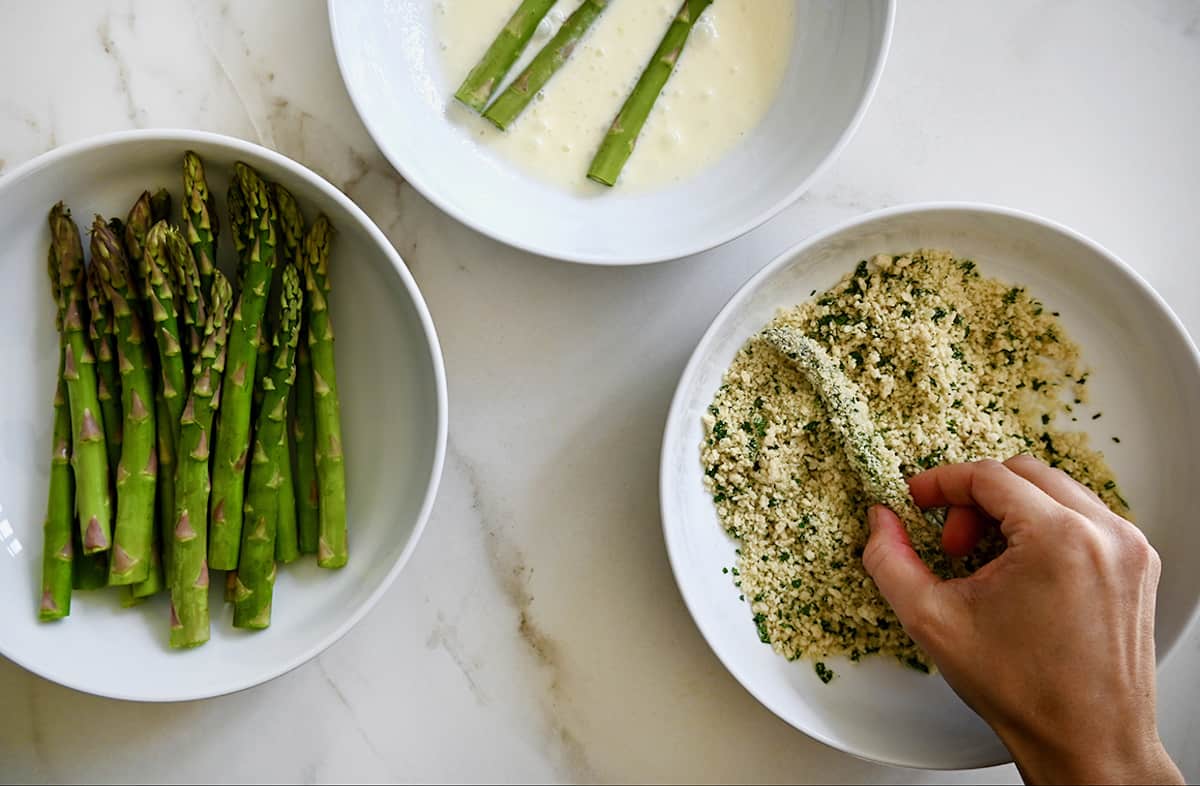 For making asparagus fries, three white bowls hold asparagus, a mayonnaise-egg white mixture, and breadcrumbs mixed with parsley and parmesan.