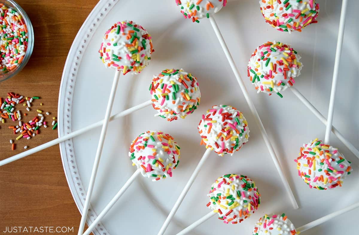 No-bake cheesecake cookie pops coated in white chocolate and rainbow sprinkles on a large white plate. A small bowl with more rainbow sprinkles is beside the plate.