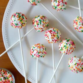 Cheesecake cookie pops coated in white chocolate and covered in rainbow sprinkles on a large white plate. A small bowl with more rainbow sprinkles is to the bottom left of the plate.