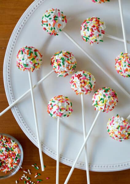 Cheesecake cookie pops coated in white chocolate and covered in rainbow sprinkles on a large white plate. A small bowl with more rainbow sprinkles is to the bottom left of the plate.