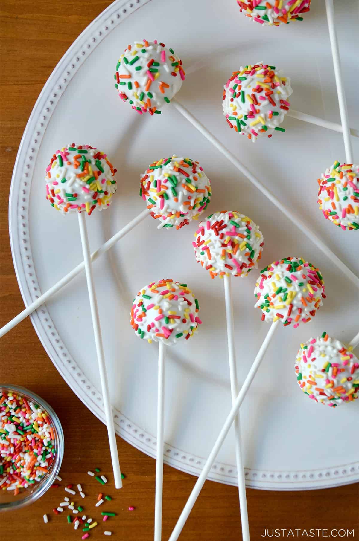 Cheesecake cookie pops coated in white chocolate and covered in rainbow sprinkles on a large white plate. A small bowl with more rainbow sprinkles is to the bottom left of the plate.