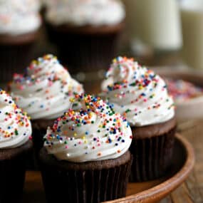 Chocolate cupcakes topped with vanilla frosting and rainbow sprinkles on a wood serving plate.