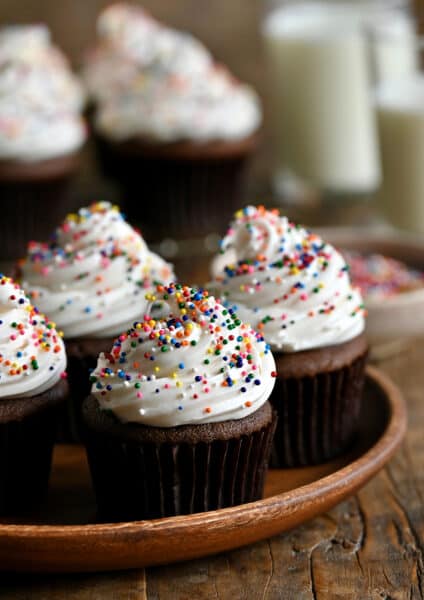 Chocolate cupcakes topped with vanilla frosting and rainbow sprinkles on a wood serving plate.