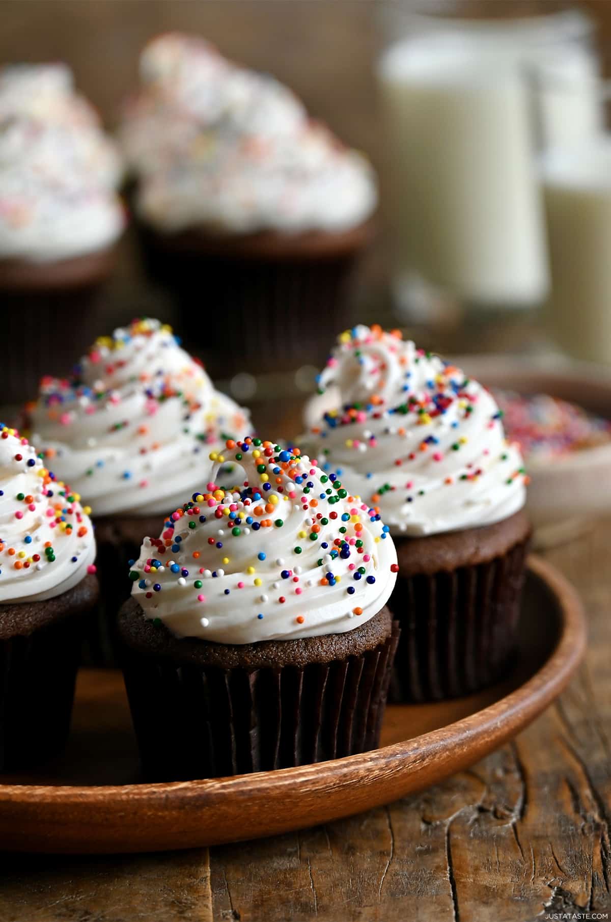 Chocolate cupcakes topped with vanilla frosting and rainbow sprinkles on a wood serving plate.