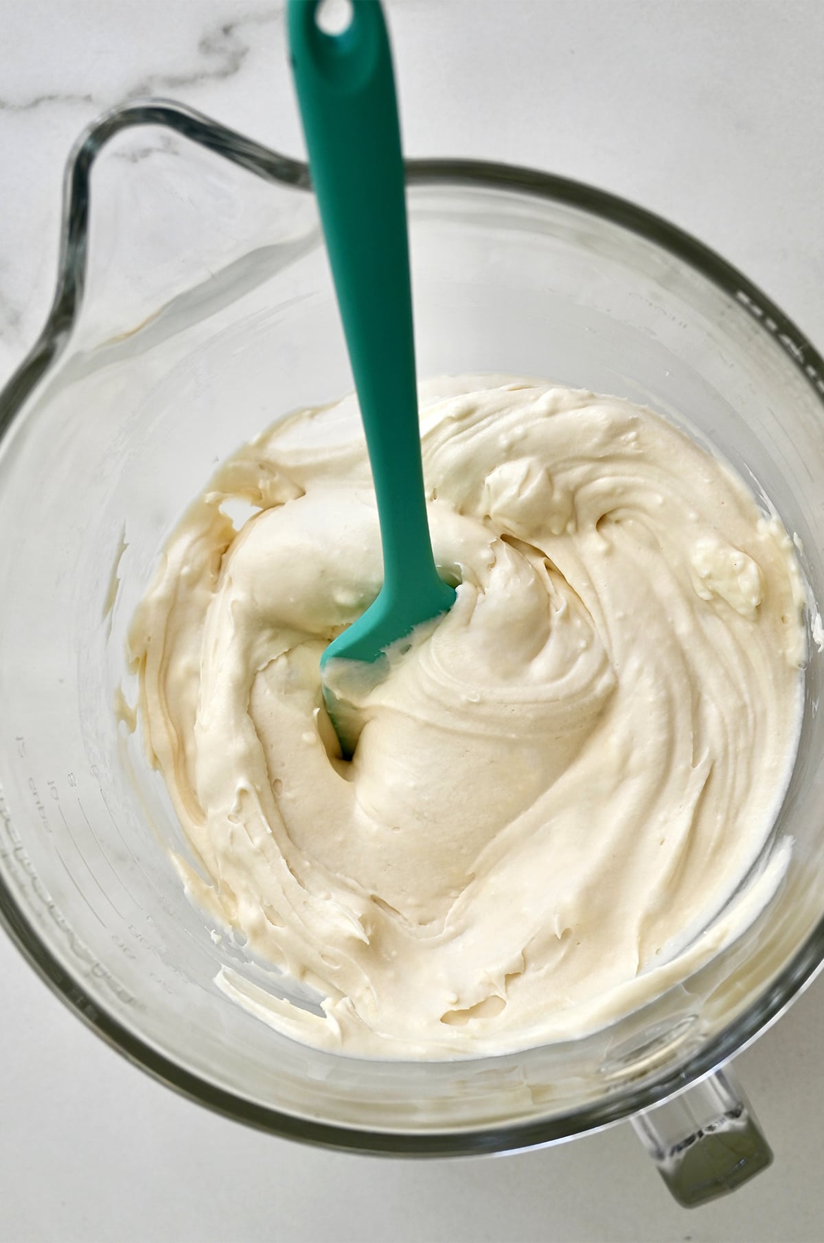 Fluffy cream cheese frosting is in a glass stand mixer bowl, with a teal spatula.