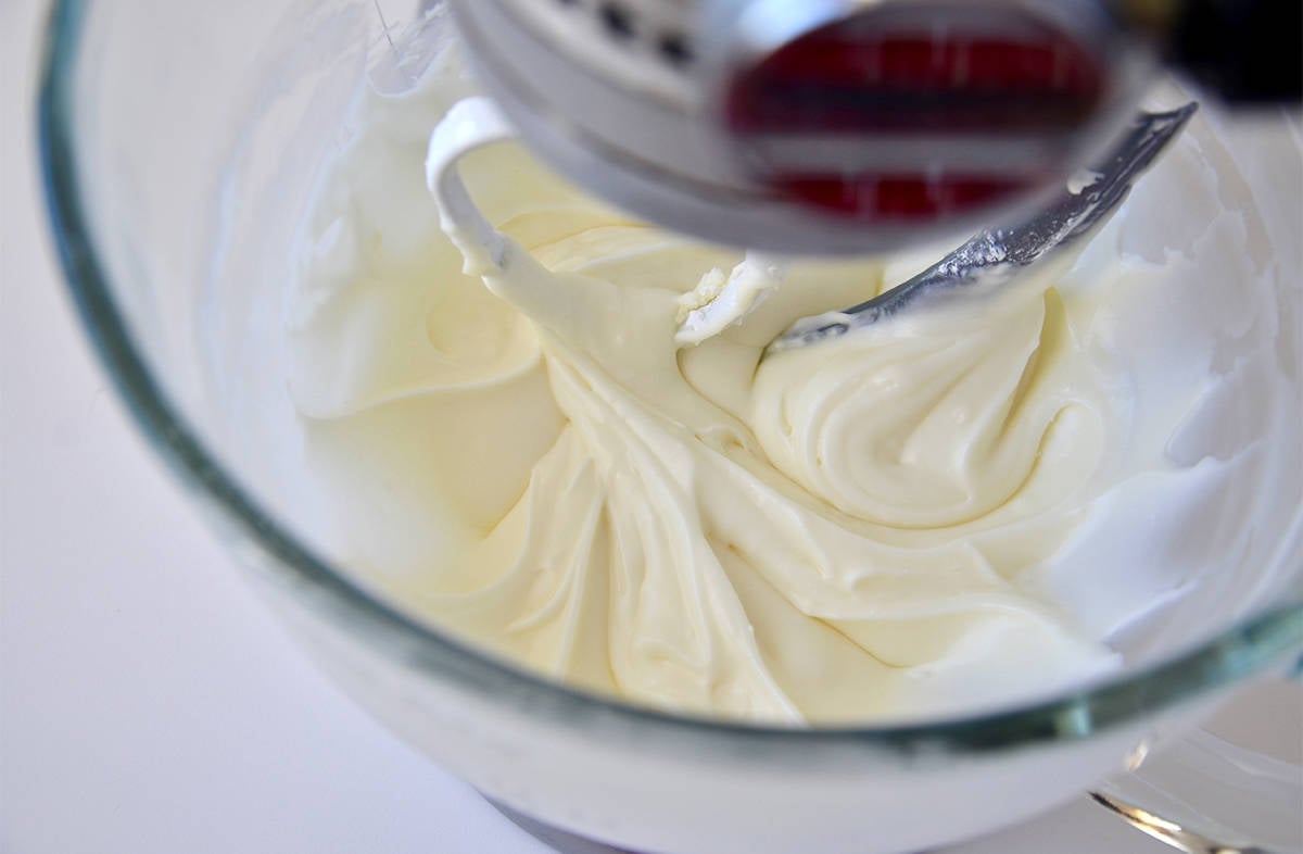 In a clear glass stand mixer bowl, cream cheese glaze is being mixed with the paddle attachment.