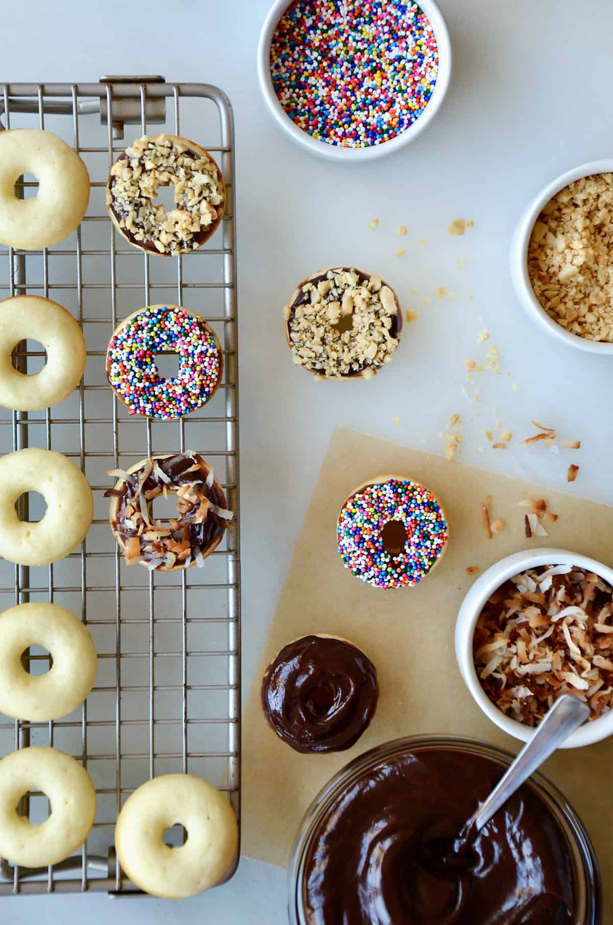 Baked doughnuts on a wire cooling rack. Topped doughnuts and bowls of Nutella glaze, sprinkles, chopped nuts and toasted coconut are beside the cooling rack.