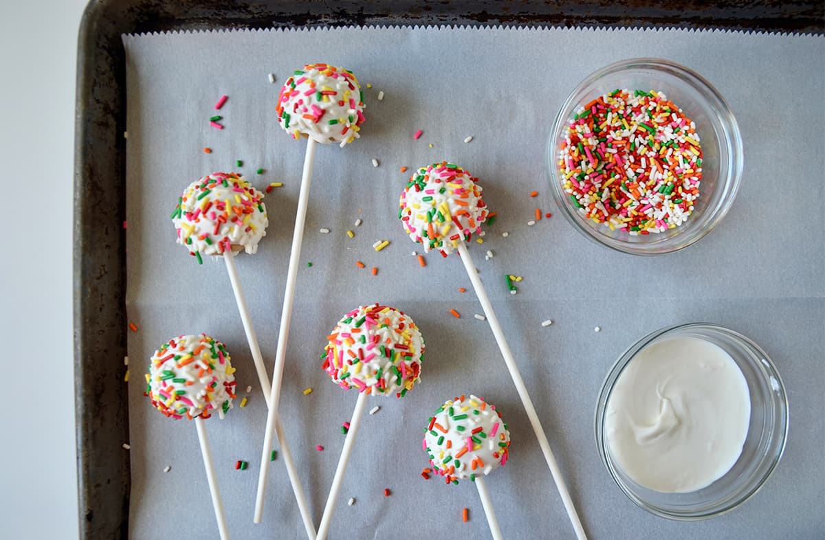 Cheesecake cookie pops covered in white chocolate and coated with rainbow sprinkles on a parchment-lined baking sheet. Small bowls of melted white chocolate and rainbow sprinkles sit beside the pops.