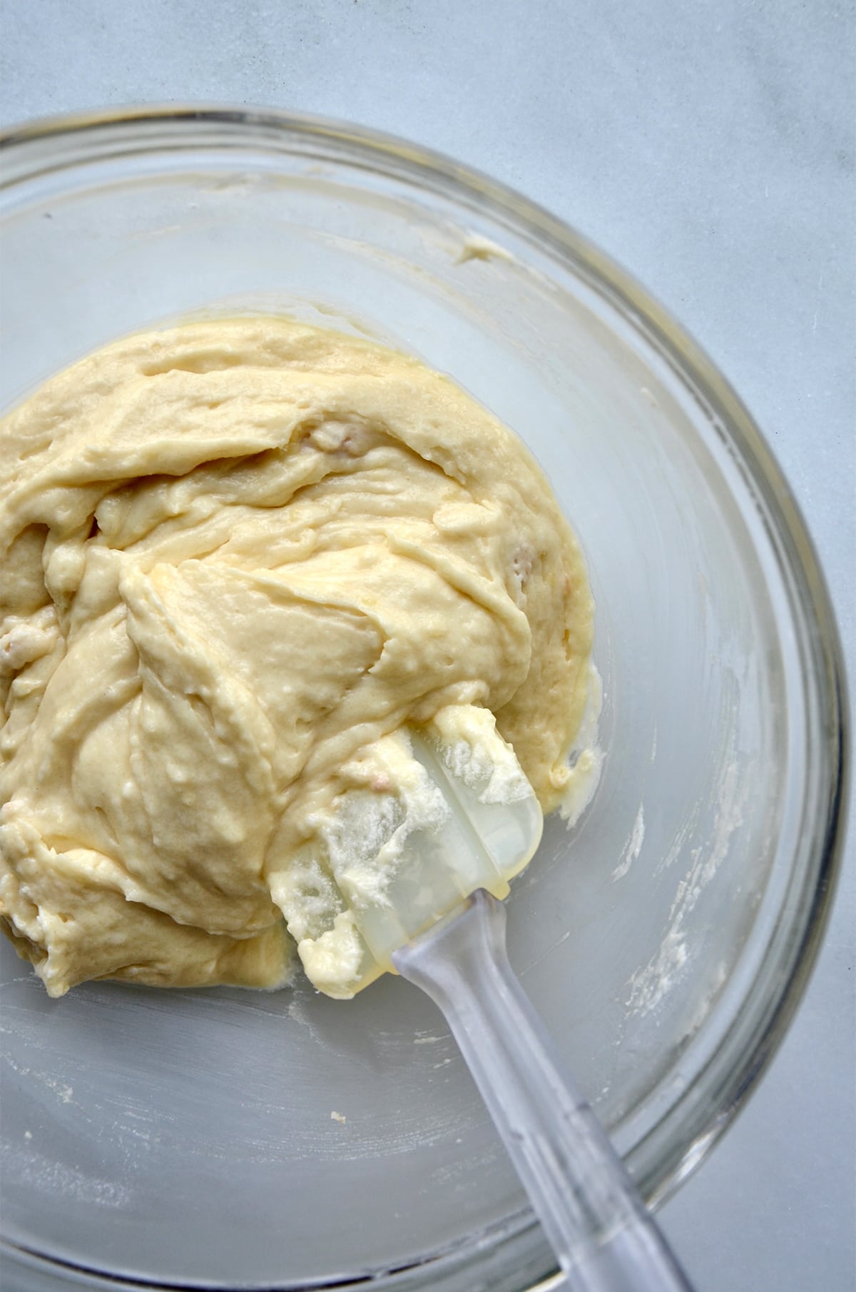 A large glass mixing bowl with doughnut batter in it. A spatula rests in the batter and against the side of the bowl.