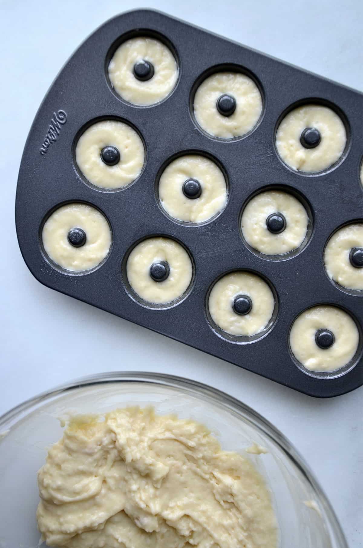 A mini doughnut pan filled with batter. A large glass mixing bowl with doughnut batter is beside the pan.