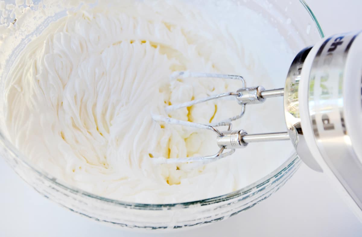 Whipped cream in a large glass mixing bowl. A hand mixer with its beaters attached rests against the side of the bowl