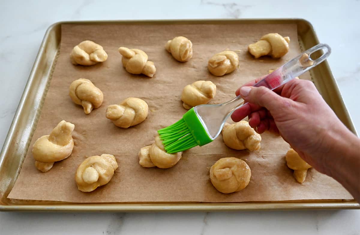 A hand holding a pastry brush coats pizza dough knots with an egg wash.