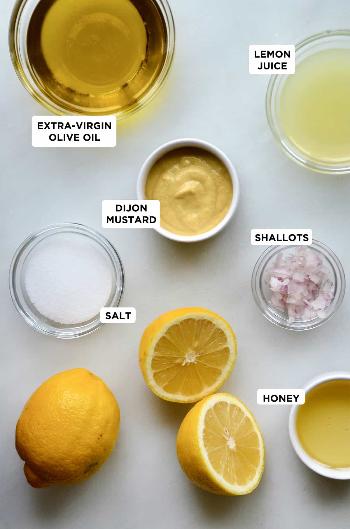 Small bowls of olive oil, mustard, salt, minced shallots, lemon juice and honey. Halved and whole lemons are beside the bowls.