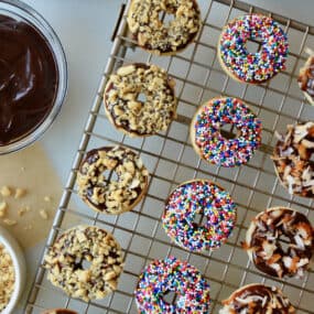 A cooling rack with baked and glazed doughnuts topped with chopped nuts, sprinkles and toasted coconut. Bowls with more toppings and Nutella glaze are beside the cooling rack.