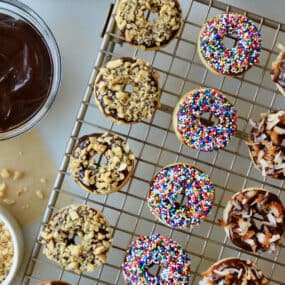 A cooling rack with baked and glazed doughnuts topped with chopped nuts, sprinkles and toasted coconut. Bowls with more toppings and Nutella glaze are beside the cooling rack.