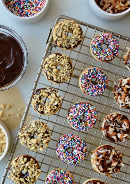 A cooling rack with baked and glazed doughnuts topped with chopped nuts, sprinkles and toasted coconut. Bowls with more toppings and Nutella glaze are beside the cooling rack.