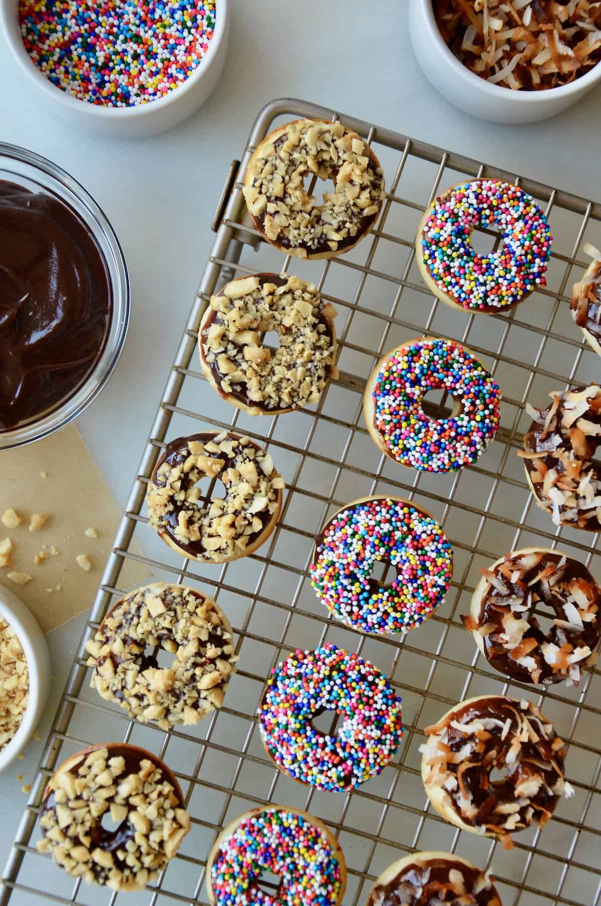 A cooling rack with baked and glazed doughnuts topped with chopped nuts, sprinkles and toasted coconut. Bowls with more toppings and Nutella glaze are beside the cooling rack.