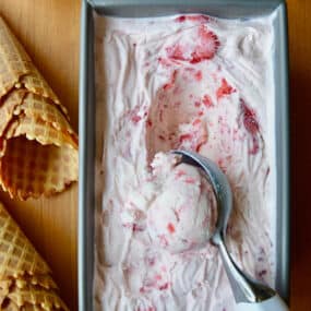 A loaf pan with no-churn strawberry ice cream and an ice cream scoop scooping out the ice cream. Waffle cones and small bowls of strawberries and fresh mint are beside the loaf pan.