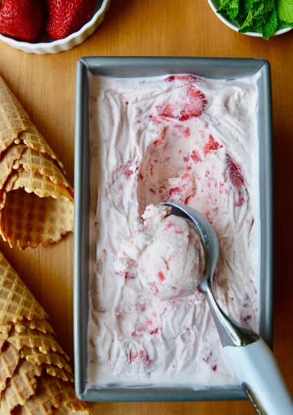 A loaf pan with no-churn strawberry ice cream and an ice cream scoop scooping out the ice cream. Waffle cones and small bowls of strawberries and fresh mint are beside the loaf pan.