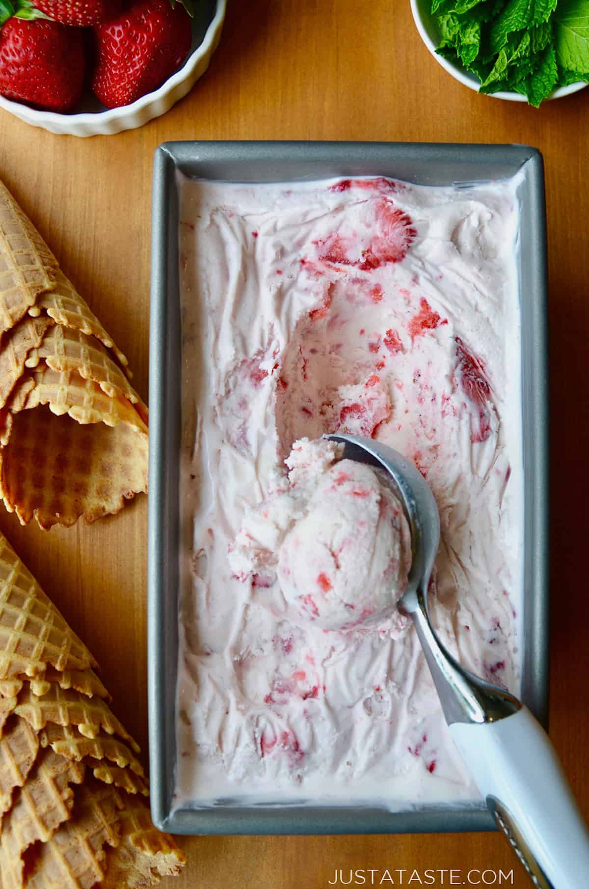 A loaf pan with no-churn strawberry ice cream and an ice cream scoop scooping out the ice cream. Waffle cones and small bowls of strawberries and fresh mint are beside the loaf pan.