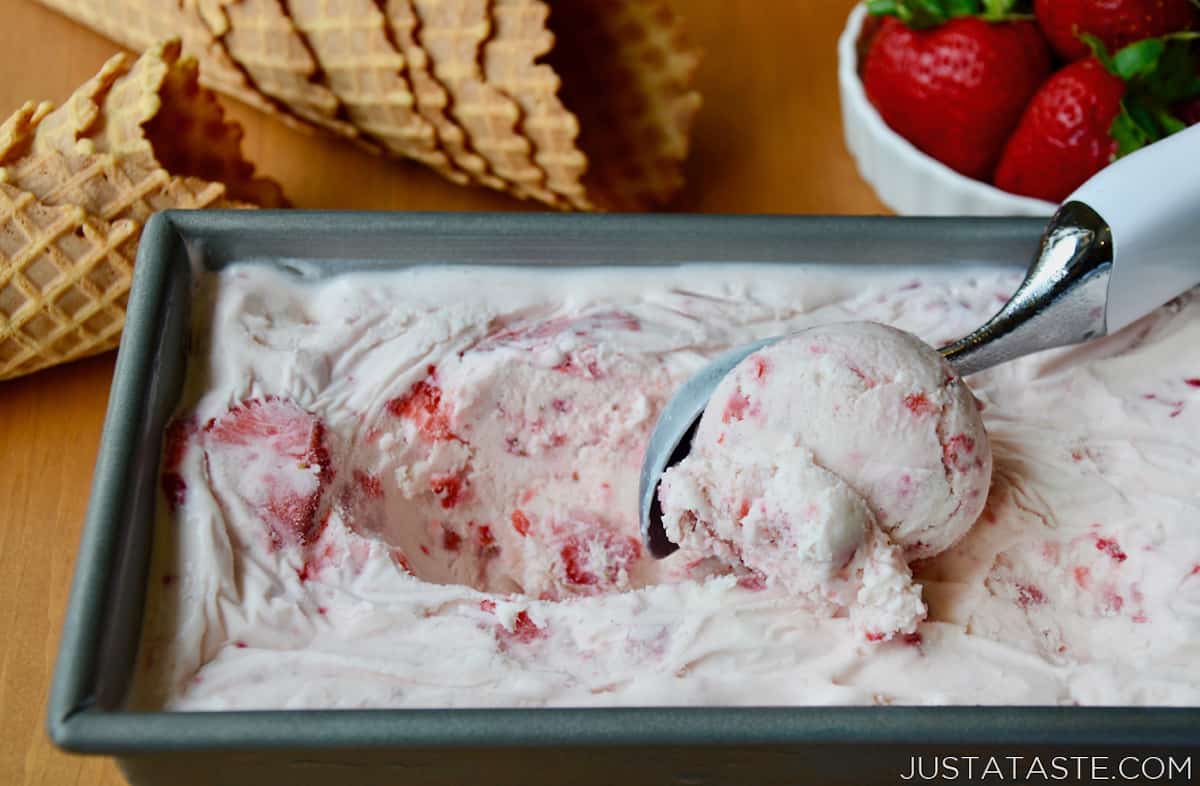 An ice cream scoop scooping no-churn strawberry ice cream out of a loaf pan. Waffle cones and fresh strawberries are next to the loaf pan.