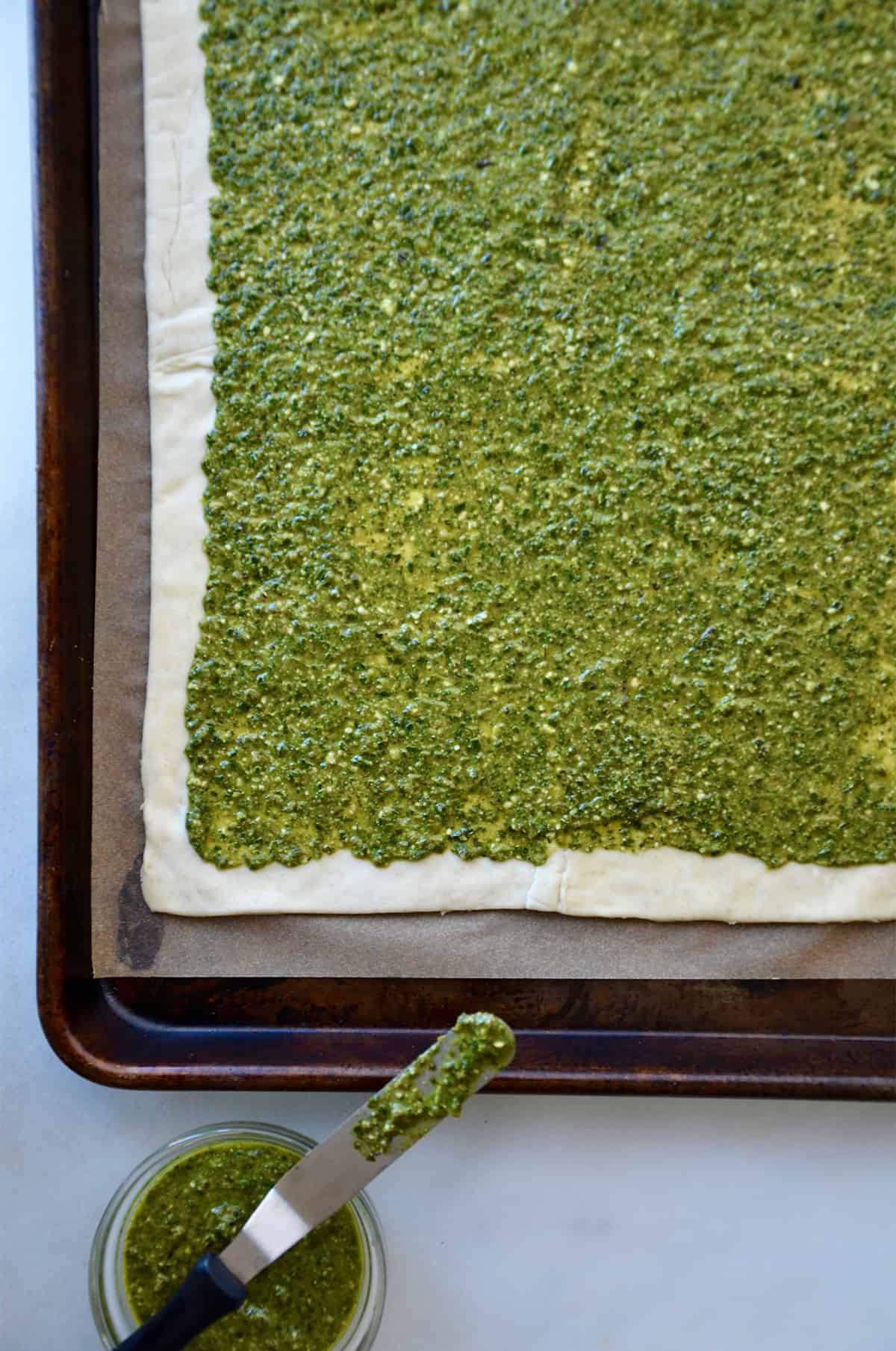 A rectangle of puff pastry topped with pesto and placed on a rimmed baking sheet lined with parchment paper.