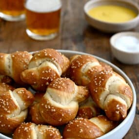 Pizza dough soft pretzel knots piled high in a white bowl with two glasses of beer nearby.