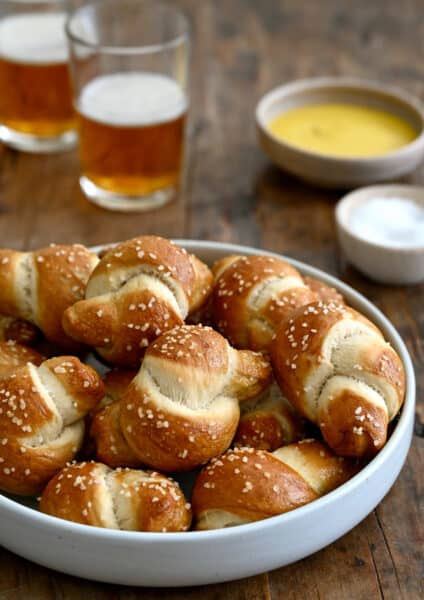 Pizza dough soft pretzel knots piled high in a white bowl with two glasses of beer nearby.