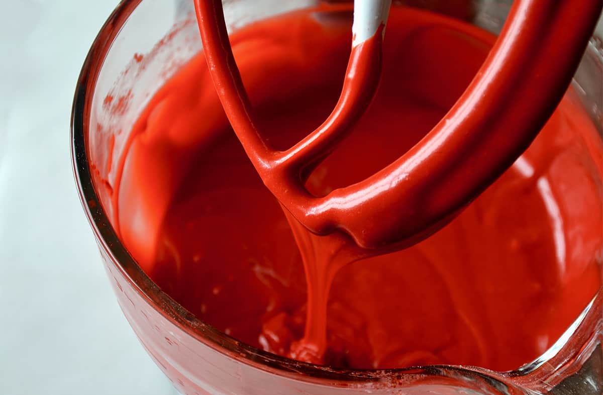 Red velvet cake batter drips from a paddle attachment into a glass stand mixer bowl.