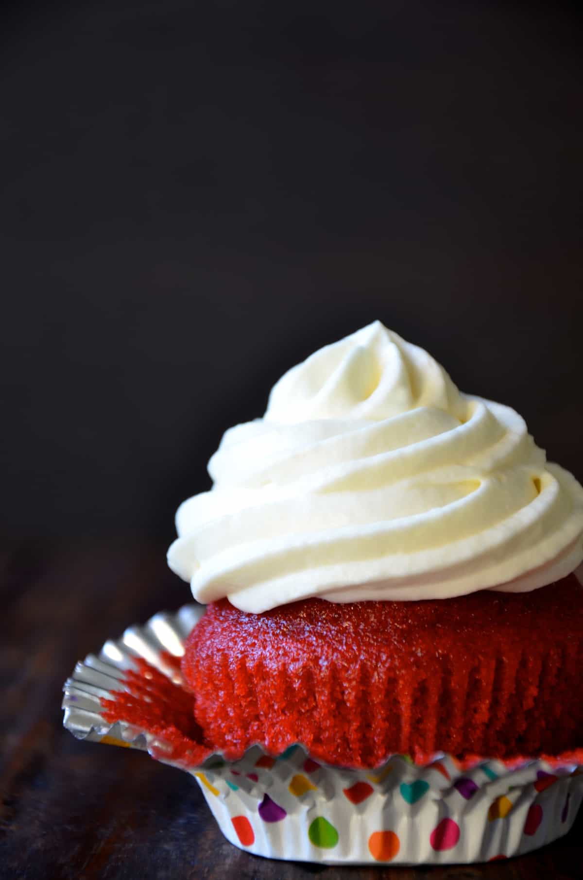 A polka-dotted cupcake paper is partially peeled away from a red velvet cupcake topped with a swirl of cream cheese frosting.