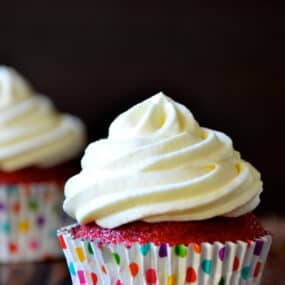 A red velvet cupcake with cream cheese frosting in a polka-dotted cupcake paper, with another cupcake in the background.