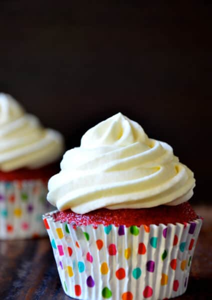 A red velvet cupcake with cream cheese frosting in a polka-dotted cupcake paper, with another cupcake in the background.