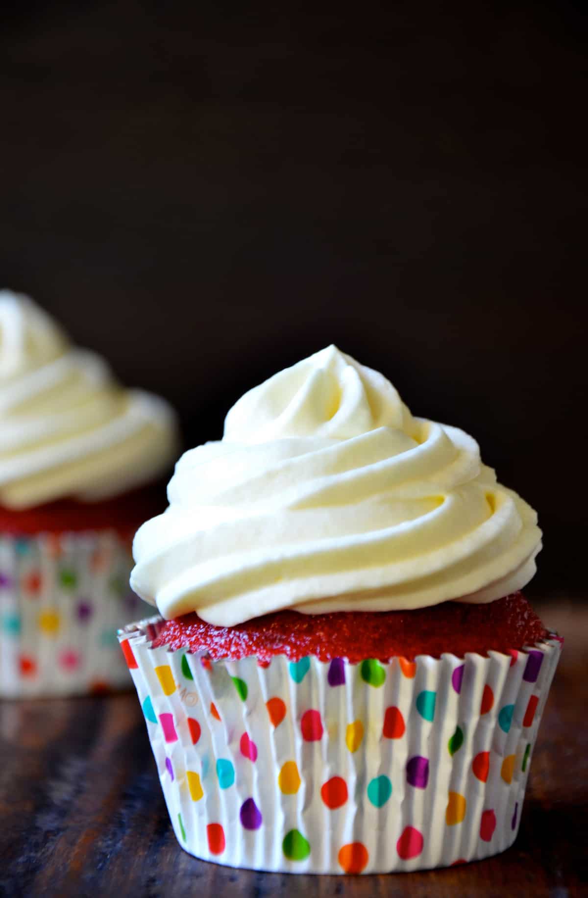 A red velvet cupcake with cream cheese frosting in a polka-dotted cupcake paper, with another cupcake in the background.