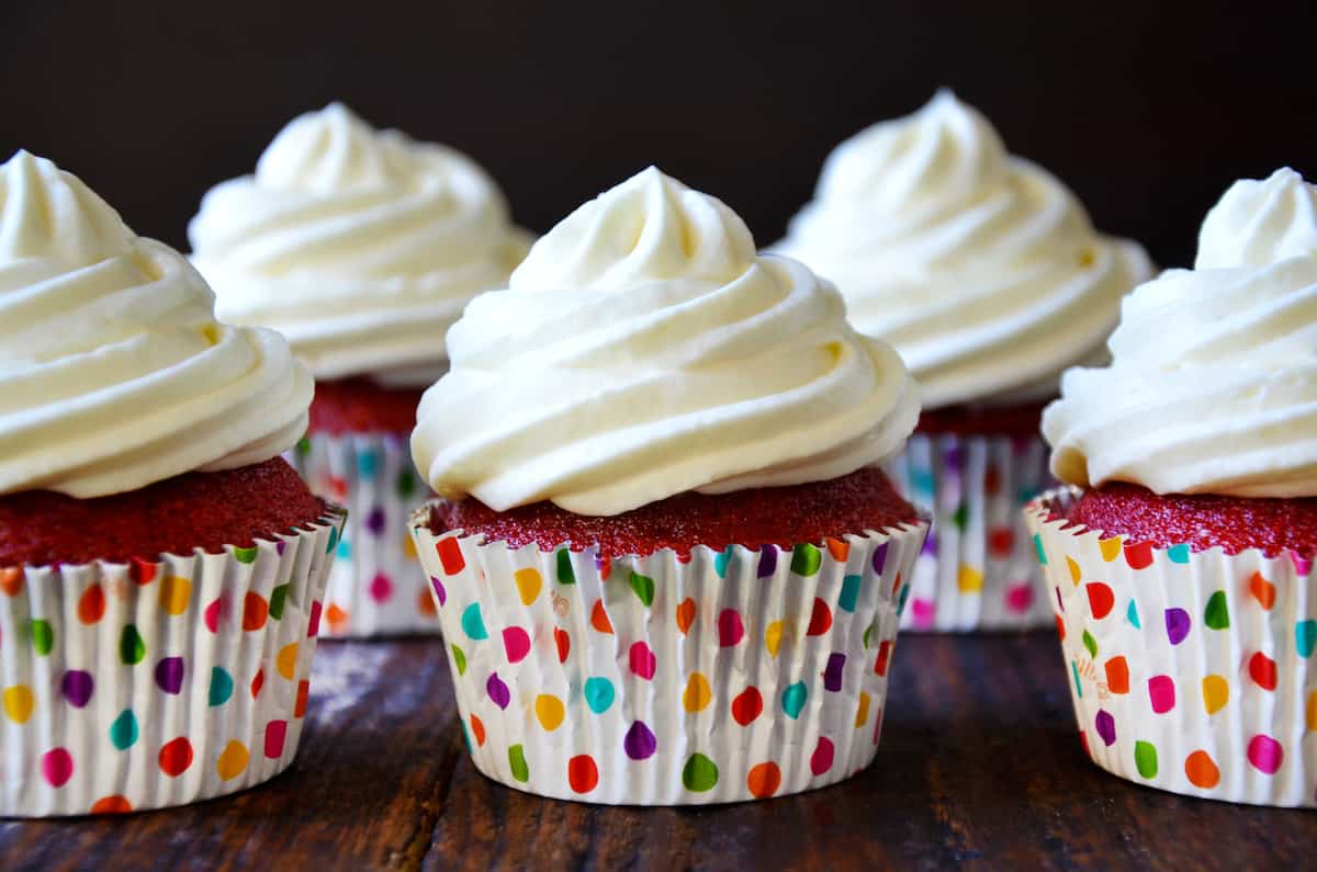 Red velvet cupcakes in polka-dotted cupcake papers, with swirls of cream cheese, are lined up in rows on a wood surface.