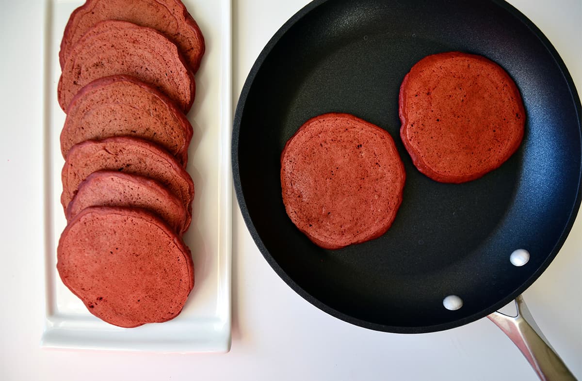 Two red velvet pancakes sit in a nonstick skillet. A stack of cooked pancakes sit nearby on a rectangular white platter.