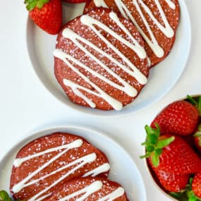Red velvet pancakes drizzled with cream cheese glaze and sprinkled with powdered sugar are on white plates and garnished with strawberries. A bowl of strawberries sits nearby.