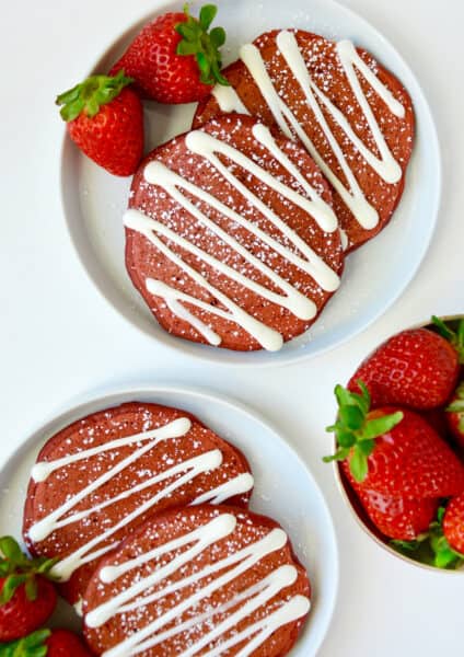 Red velvet pancakes drizzled with cream cheese glaze and sprinkled with powdered sugar are on white plates and garnished with strawberries. A bowl of strawberries sits nearby.