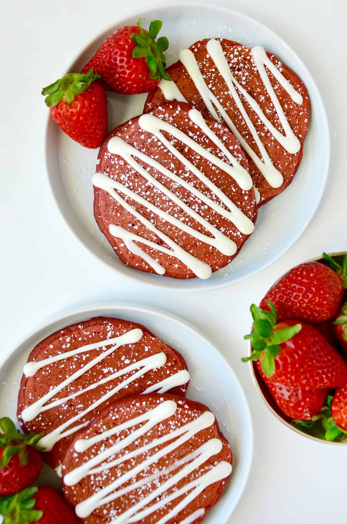Red velvet pancakes drizzled with cream cheese glaze and sprinkled with powdered sugar are on white plates and garnished with strawberries. A bowl of strawberries sits nearby.